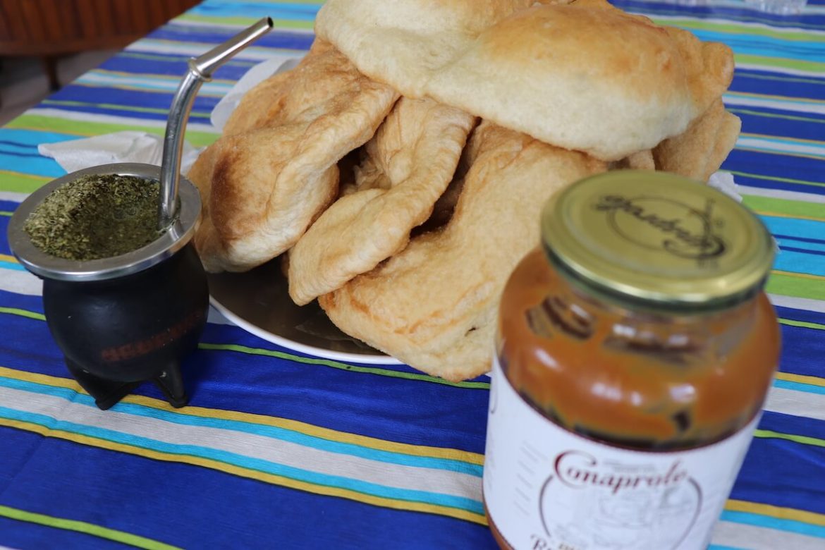 Im Vordergrund ist ein Glas Dulce de Leche zu sehen, dahinter steht eine Kalebasse mit Matetee und ein Teller mit Torta Frita auf dem Tisch